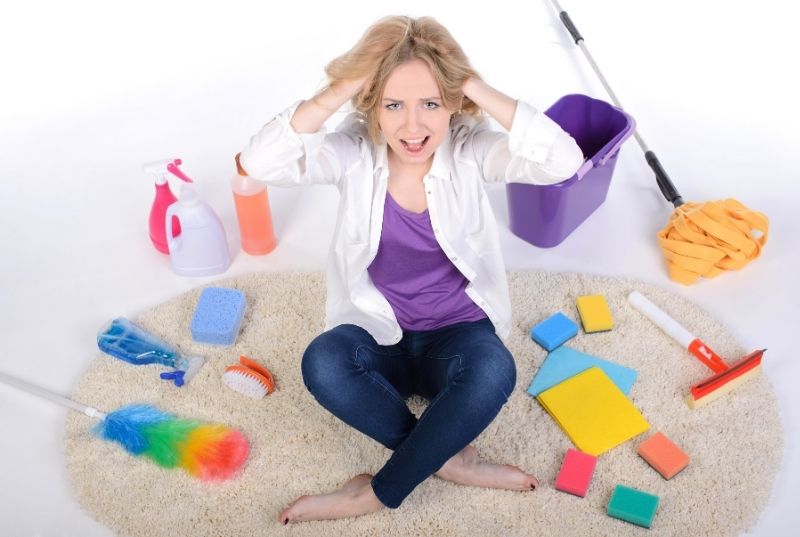 Frustrated woman sitting on a rug holding her head surranding cleaning products and tools.